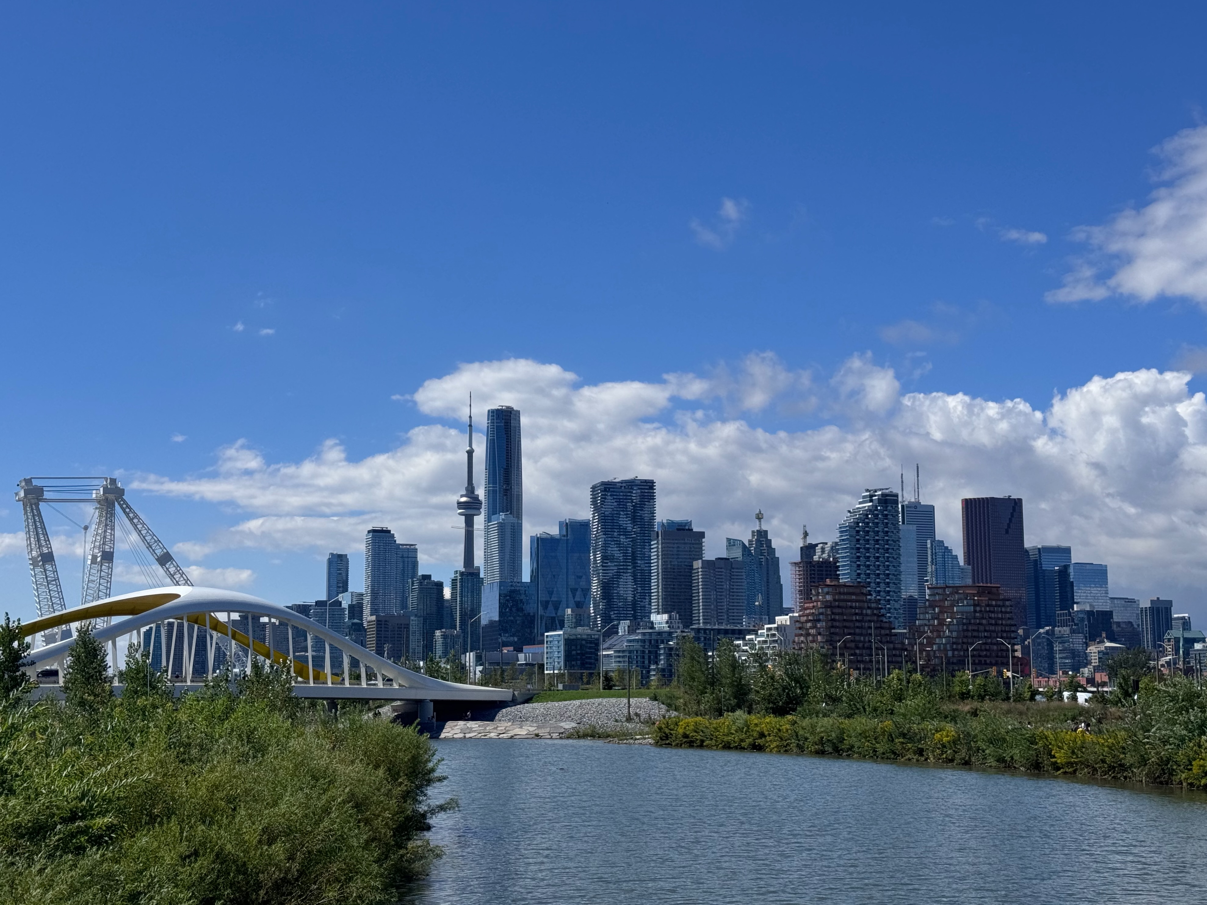 The Toronto skyline with the CN Tower rises beyond a white arched bridge over a river in a new waterfront park.