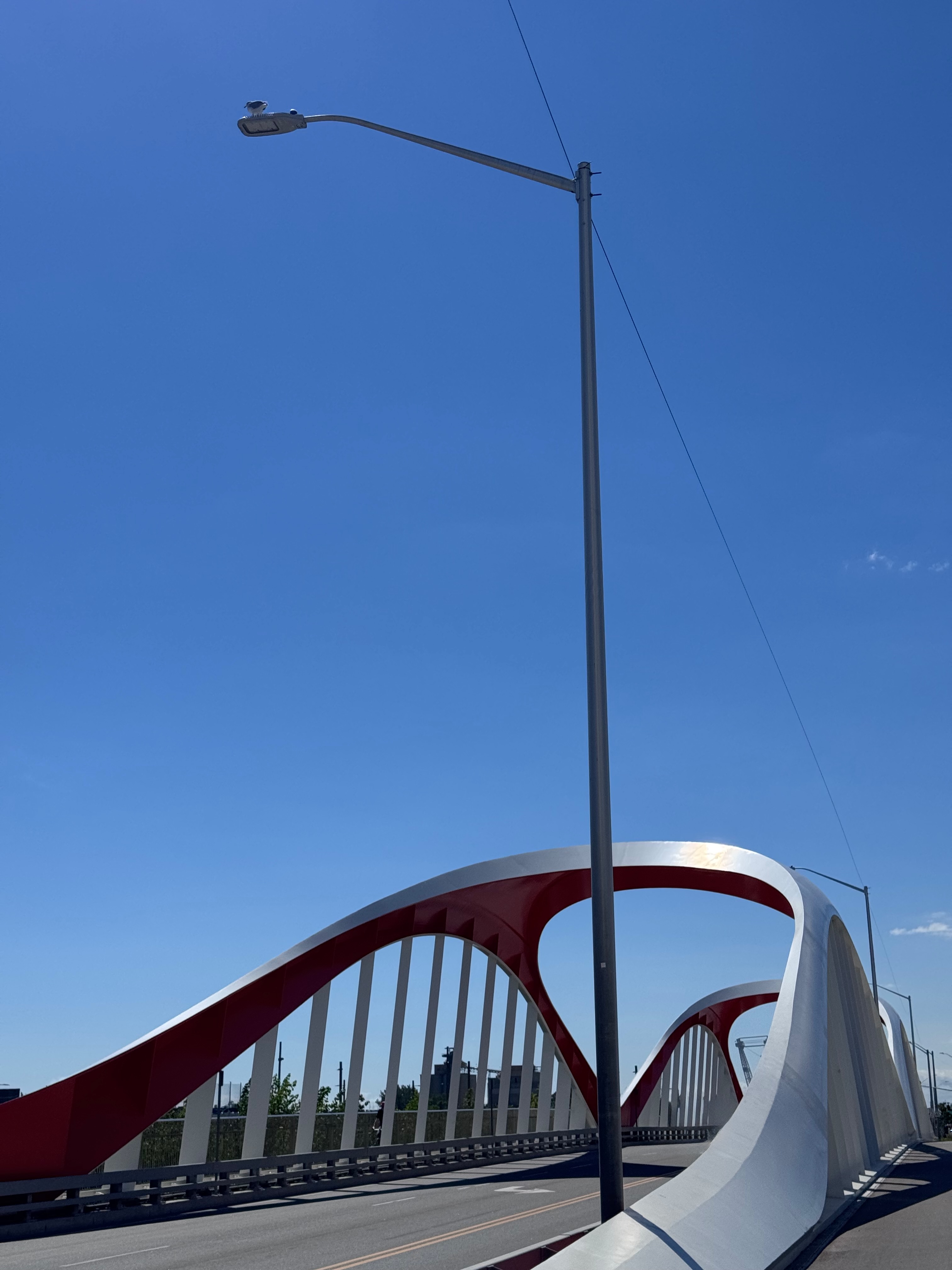 Sculptural red-and-white arched bridge with a streetlight against a clear blue sky.