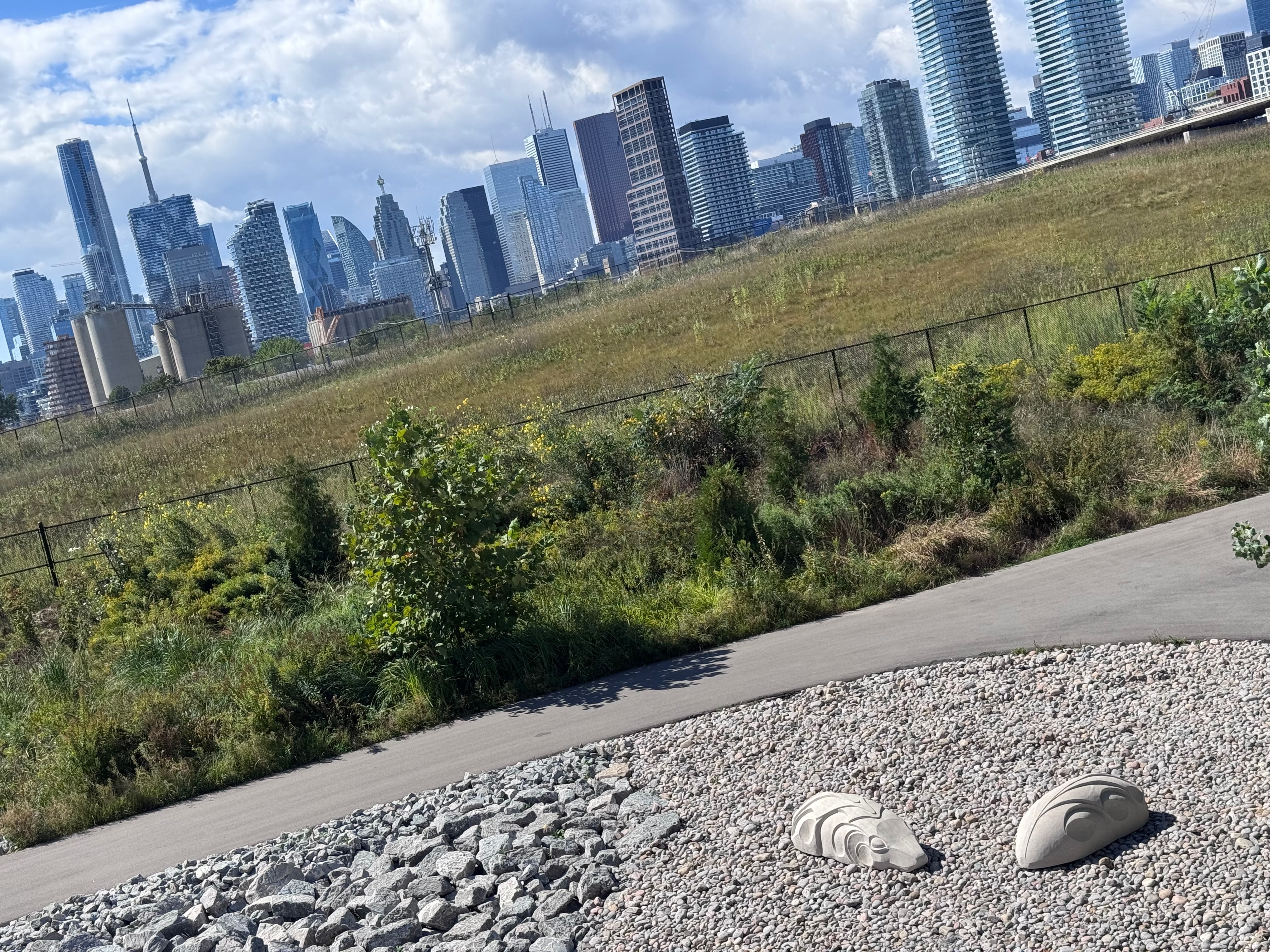 Carved stone sculptures on a gravel bed beside a meadow path, with the Toronto skyline and CN Tower behind.