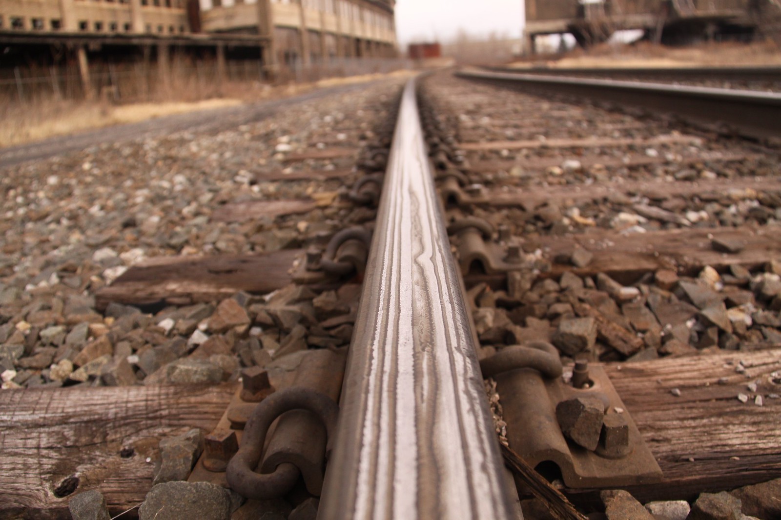 Worm's-eye view straight down a polished rail toward abandoned brick factory buildings, with rusted spikes and weathered ties.