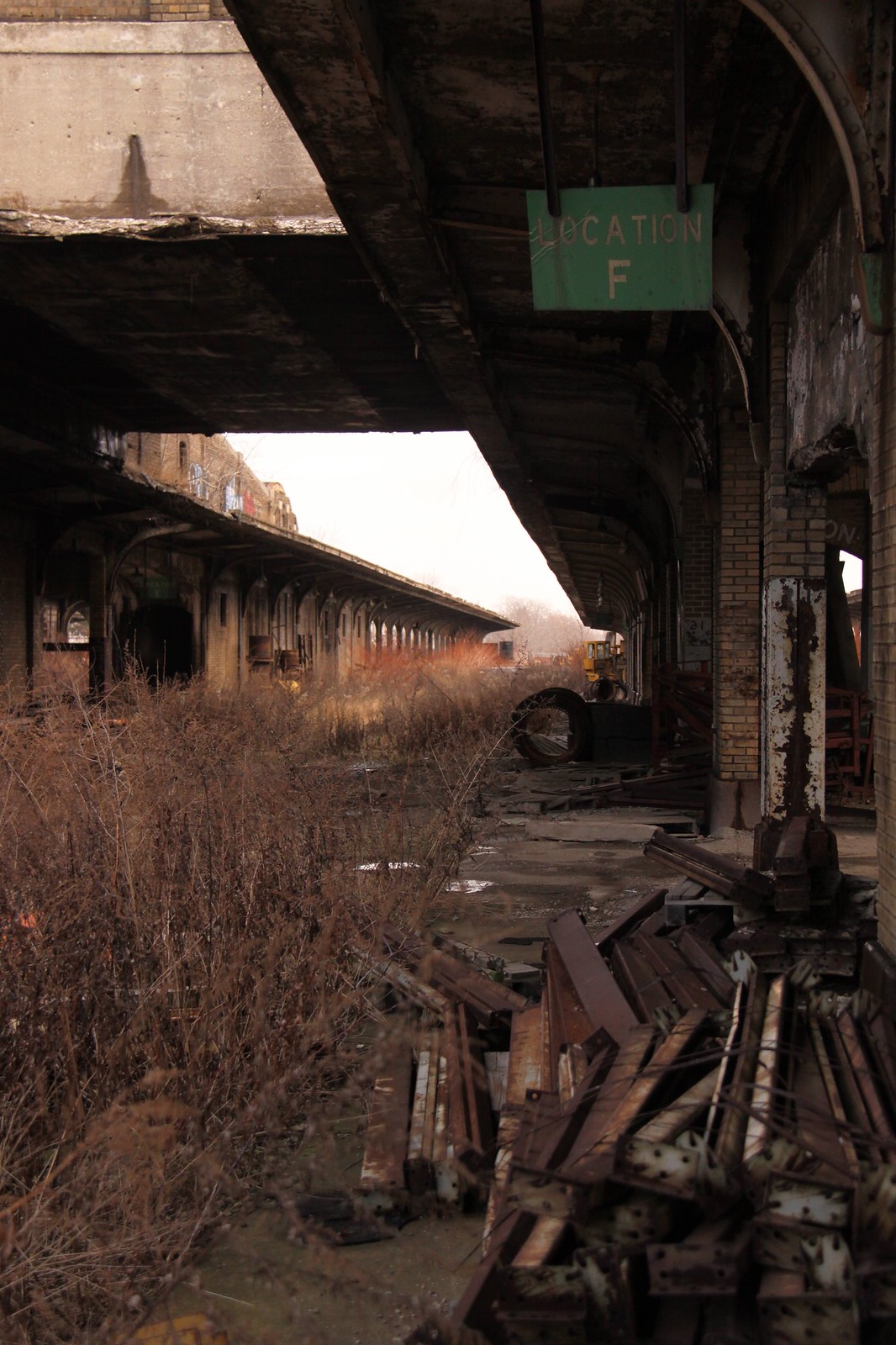 Abandoned overgrown train platform with rusted debris under a "Location F" sign.