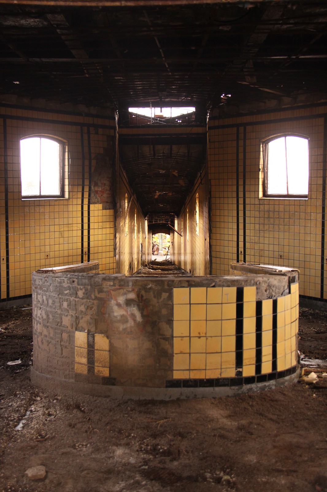 Decayed yellow-tiled room with a curved partial wall and long tunnel-like corridor in an abandoned building.