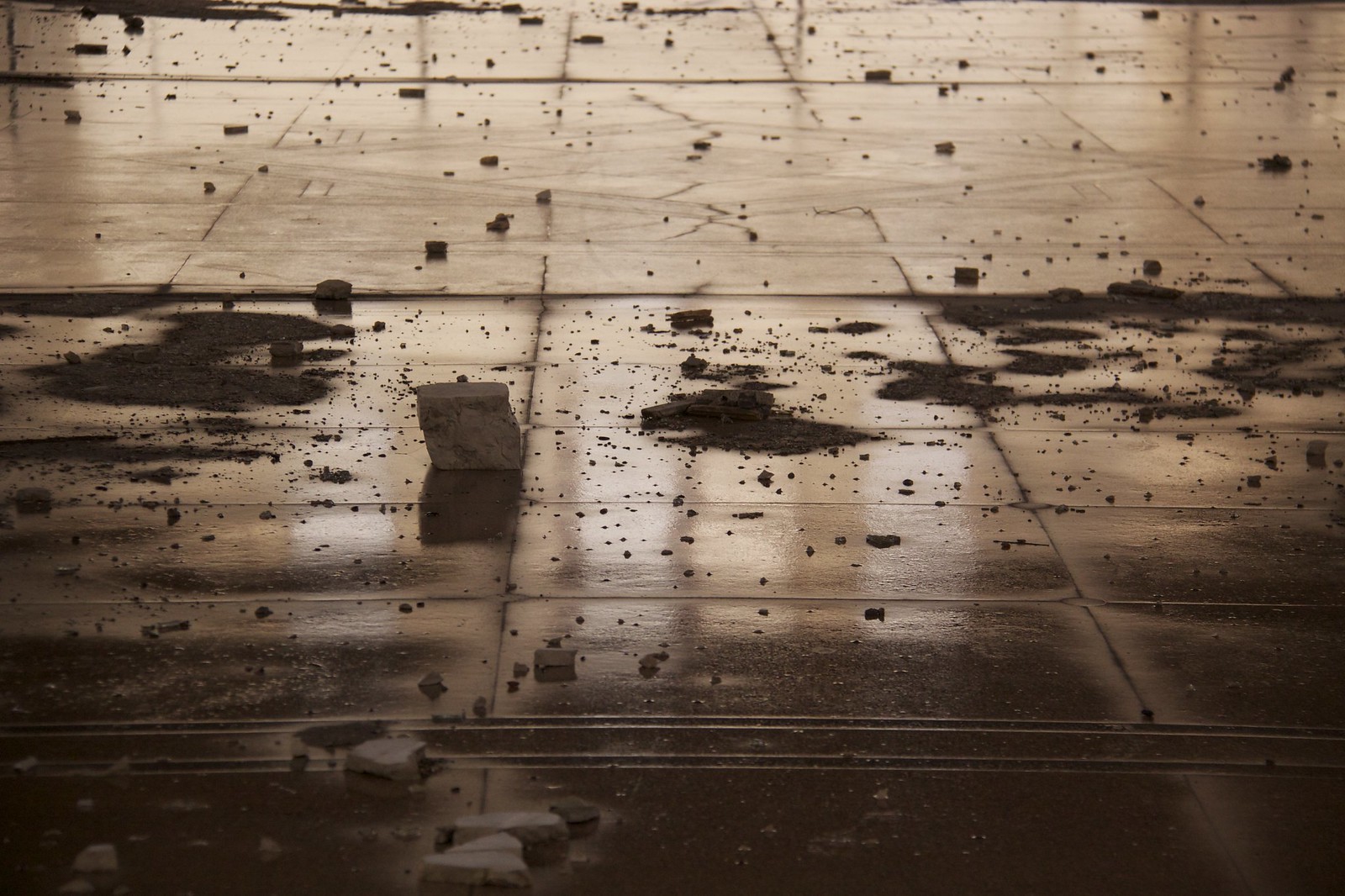 A wet, reflective tiled floor scattered with fallen debris, rubble, and dark stains in an abandoned building.
