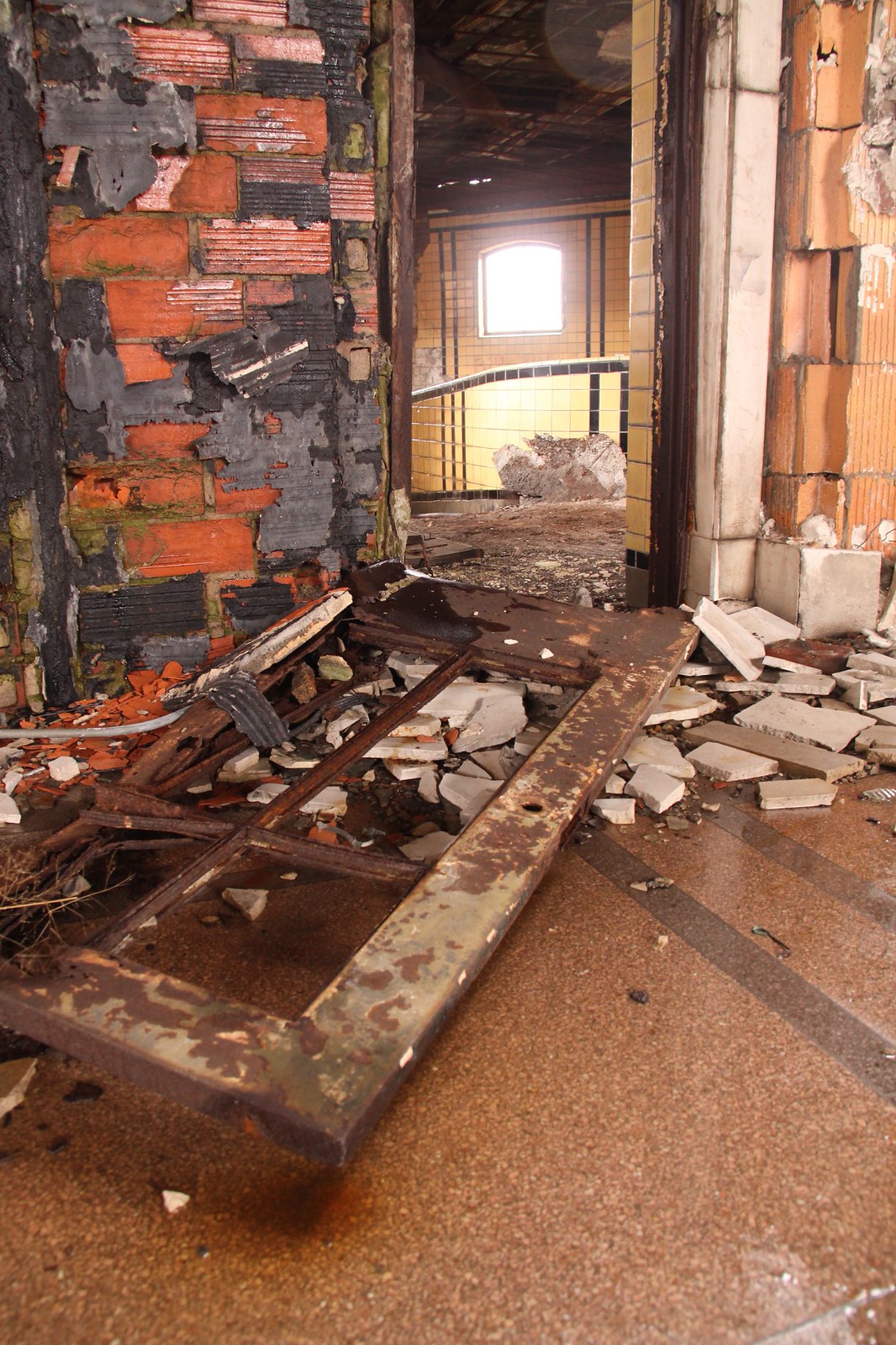 A rusted door lies fallen amid rubble in a derelict, decaying tiled room with crumbling brick walls.