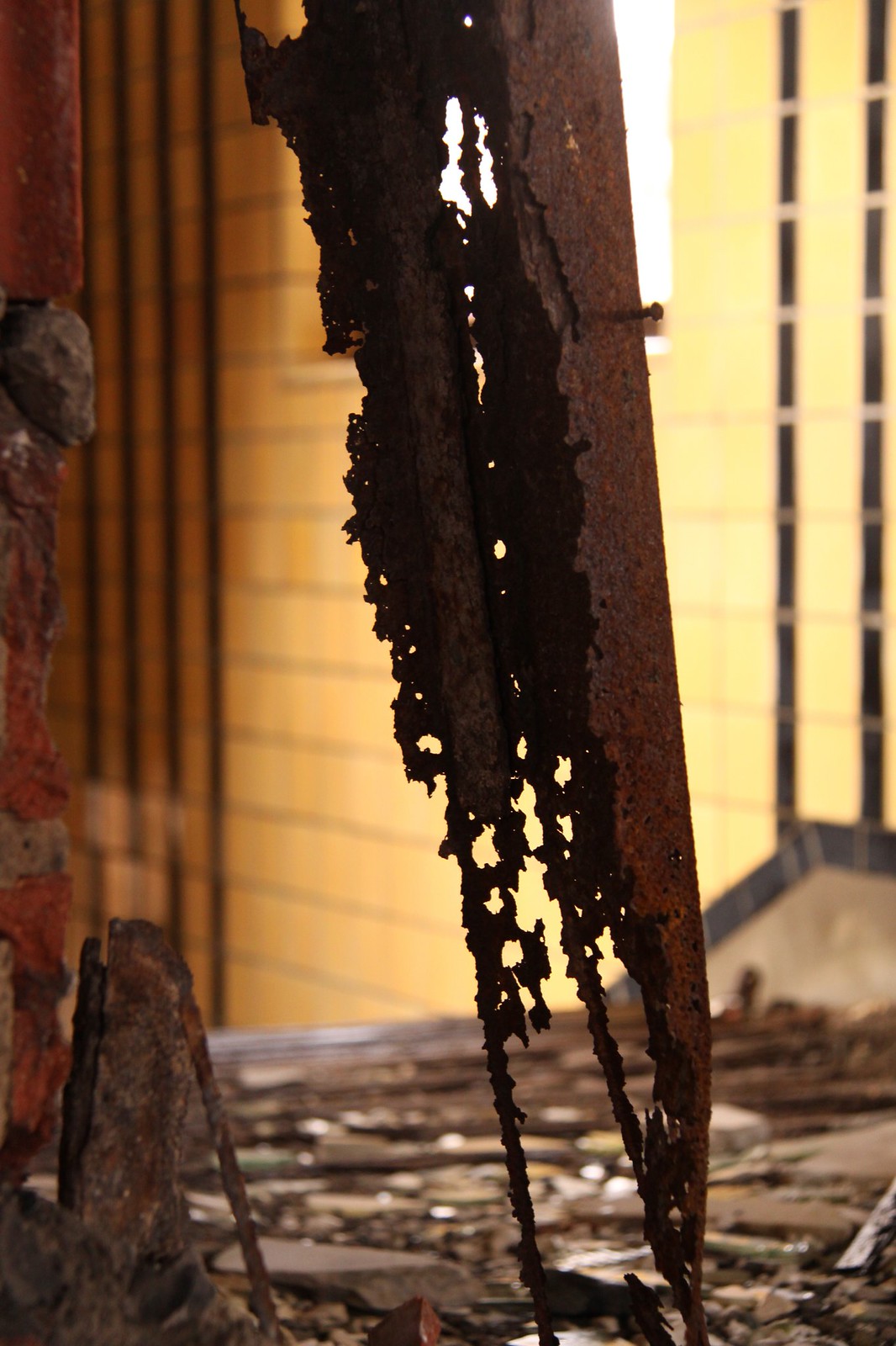 Rusted, corroded steel beam eaten through with holes against a golden tiled wall in a decaying interior.