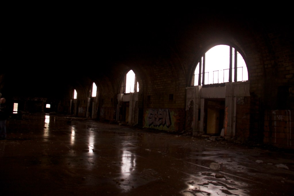 Row of tall arched windows glowing bright in a dark, abandoned graffiti-tagged hall with a wet reflective floor.