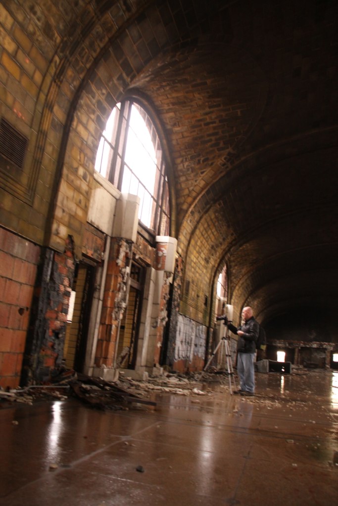 A photographer with a tripod stands in a decaying, vaulted brick hall lit by a tall arched window.