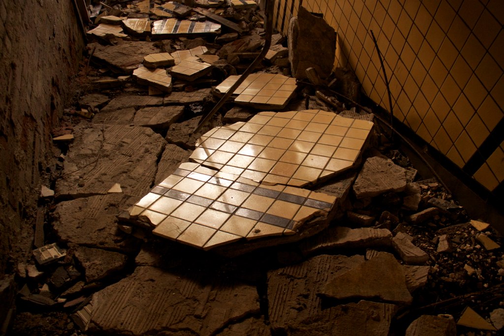 Cracked and crumbling tan tile floor amid rubble in a decaying, abandoned interior corner.