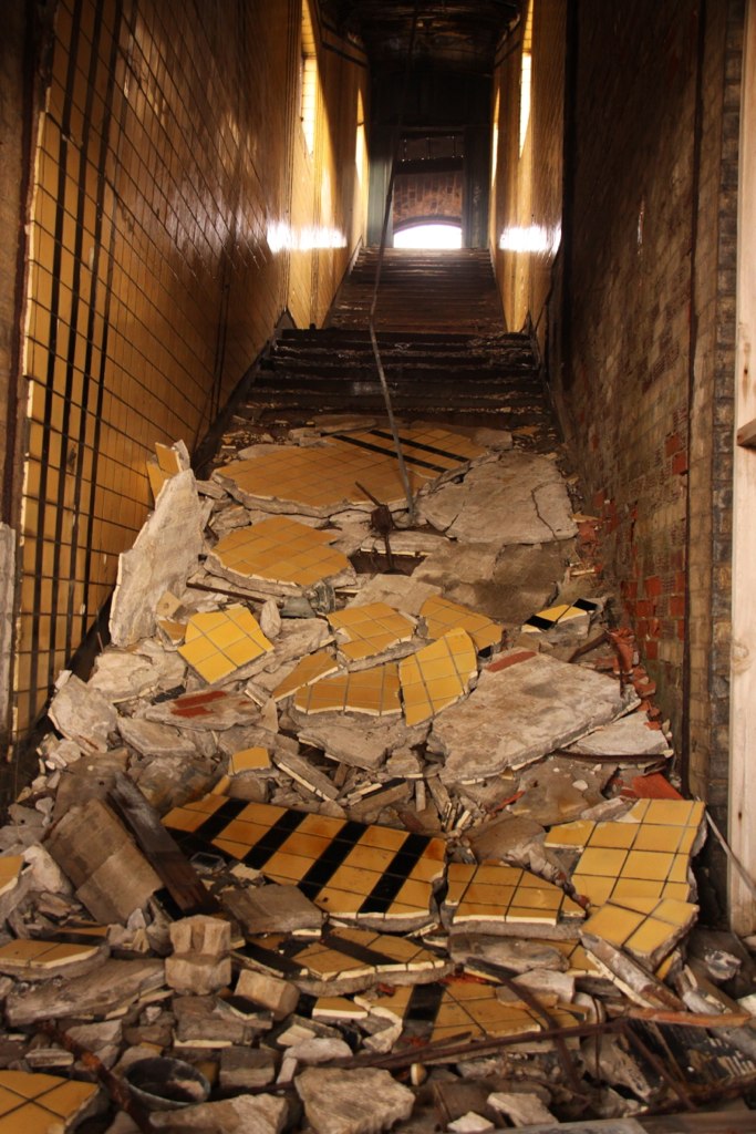 Crumbling yellow tiles and plaster debris piled at the foot of a decayed staircase in an abandoned building.