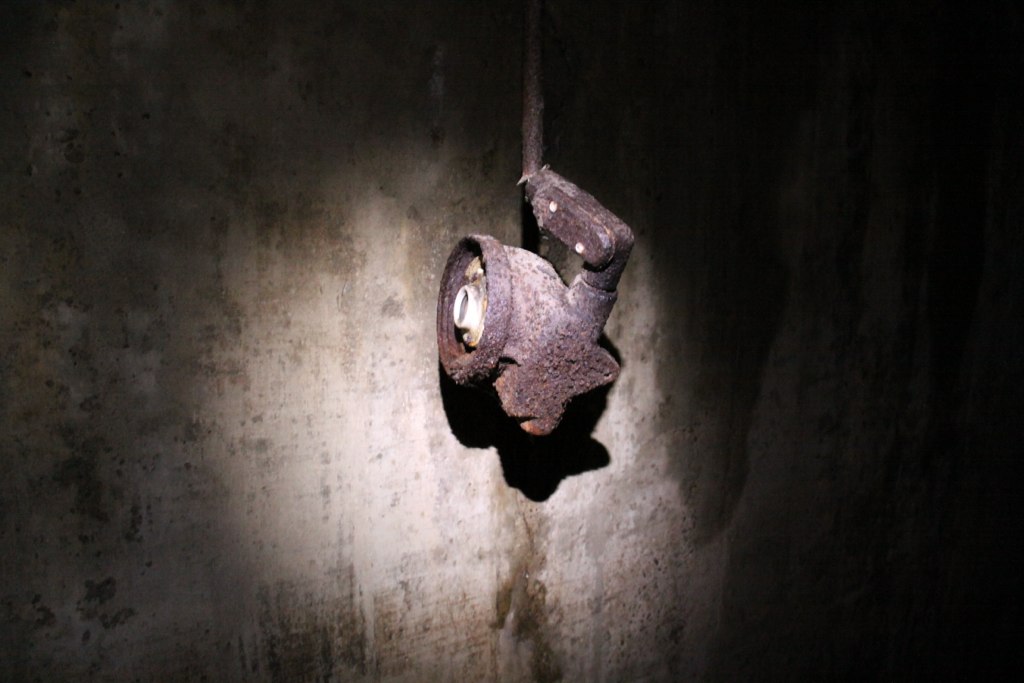 A rusted industrial light fixture juts from a dark concrete wall, its beam glowing against the grime.