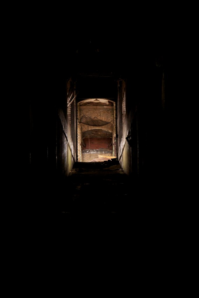 View down a dark tunnel toward a dimly lit arched brick chamber in an abandoned building