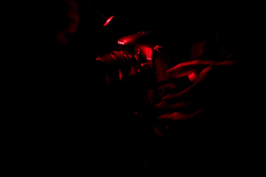 A person lit by red light holds a Canon EOS DSLR up to their eye in near-total darkness.