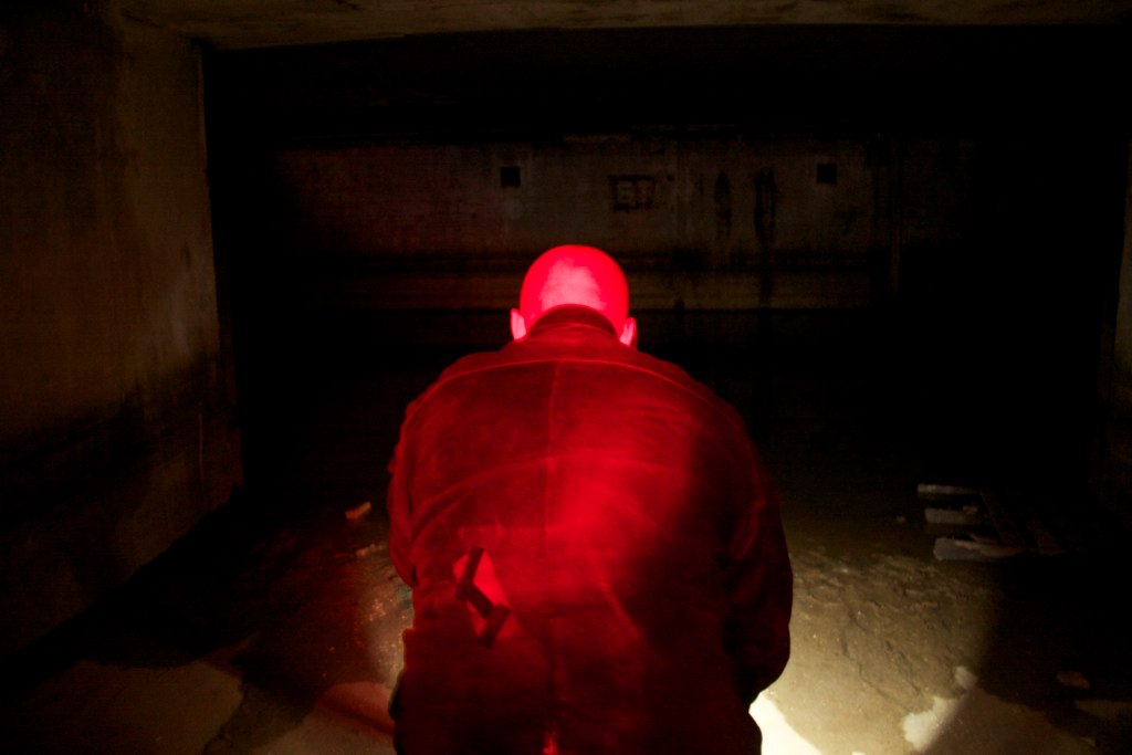 Person bathed in red light seen from behind in a dark, derelict concrete basement room.