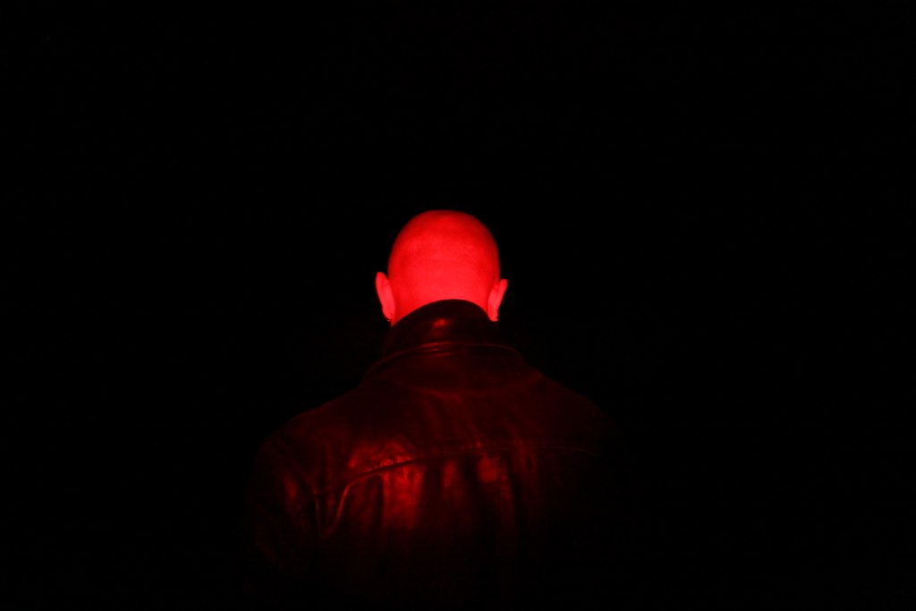 Bald man seen from behind, lit only by deep red light against a black background.