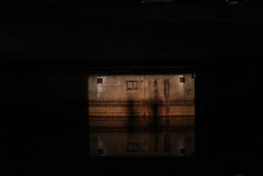 Sunlight falls on a weathered concrete wall through a dark opening, casting long shadows in a derelict interior.