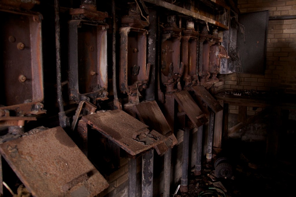 Row of rusted, decaying high-voltage electrical switch panels in an abandoned brick room.
