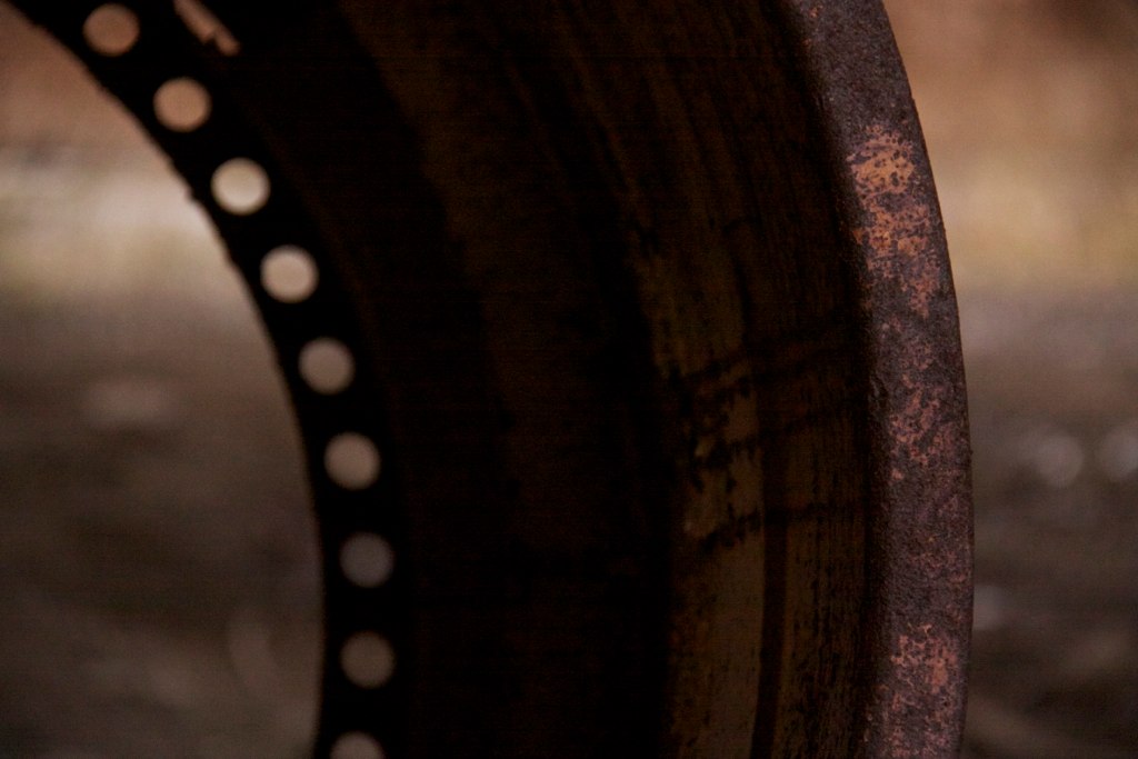Rusted metal rim of an old railcar wheel, with bolt holes curving along its inner edge, in dim light.