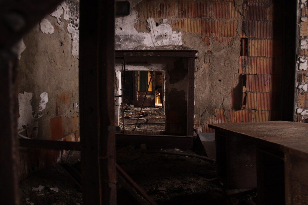 View through a doorway into decaying rooms with crumbling brick walls, exposed plaster, and rusted debris.