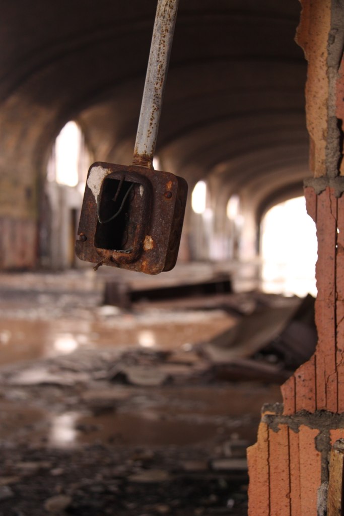 Rusted electrical switch and exposed brick in a decaying, arched, debris-strewn corridor of an abandoned building.