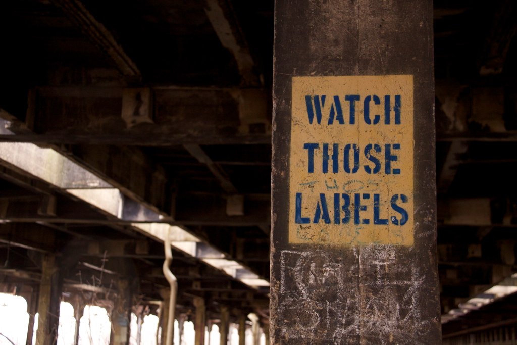 Weathered concrete pillar in an abandoned industrial hall stenciled "WATCH THOSE LABELS" in blue.