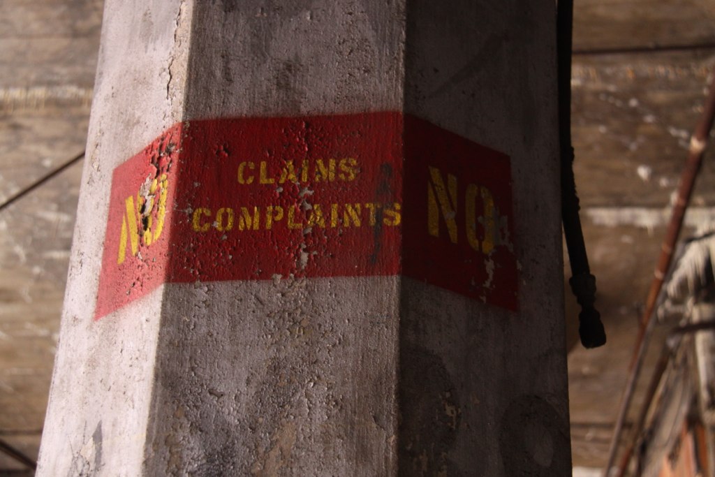 Weathered concrete pillar with a faded red band stenciled "NO CLAIMS NO COMPLAINTS" in yellow.