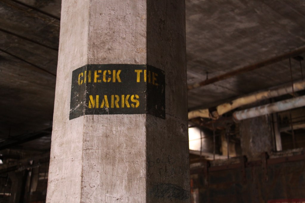 Concrete pillar in a derelict industrial basement stenciled "CHECK THE MARKS" in yellow.