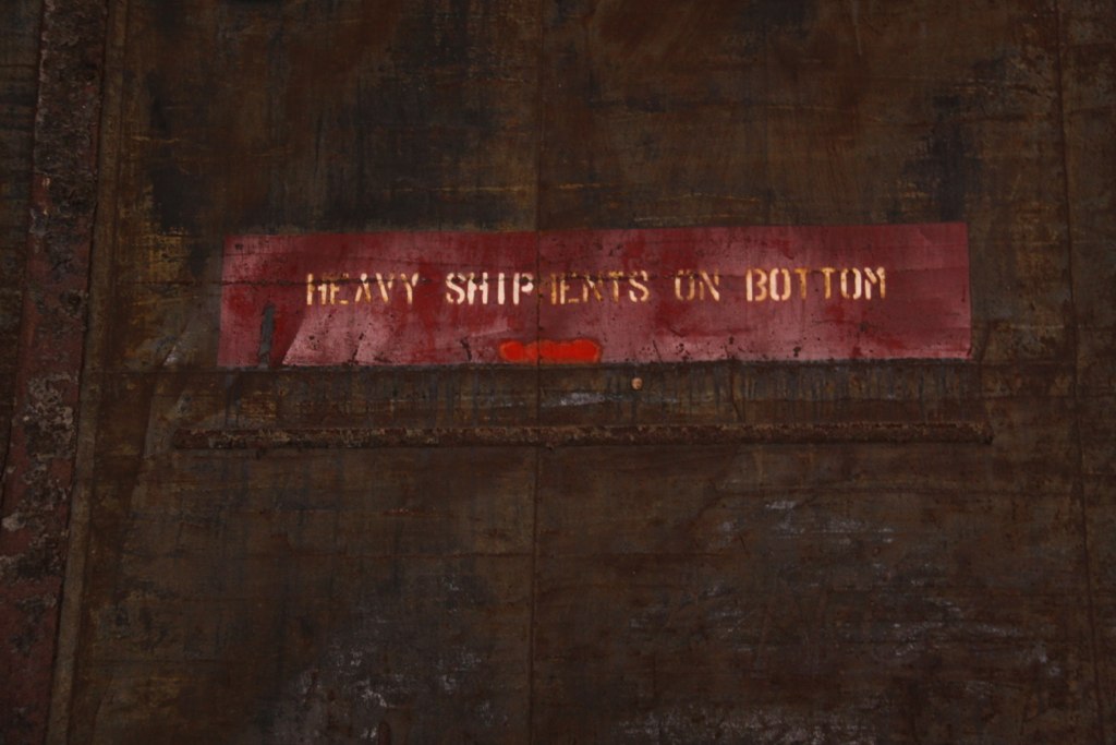 Faded red stencil reading "HEAVY SHIPMENTS ON BOTTOM" on a weathered, rusted metal wall.