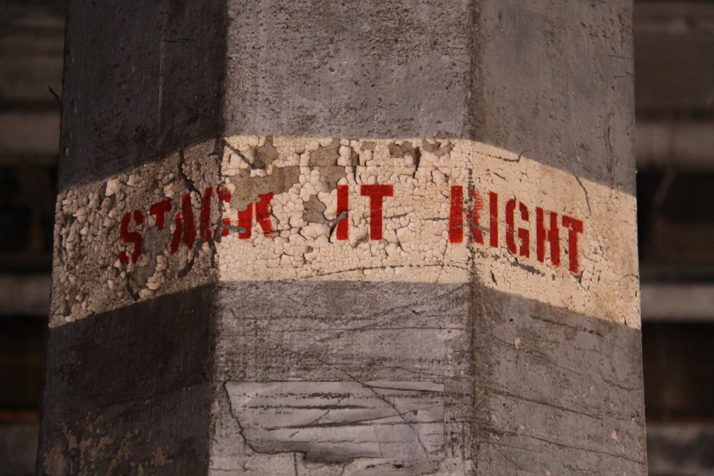 Weathered concrete pillar with peeling paint stenciled "STACK IT RIGHT" in faded red letters.