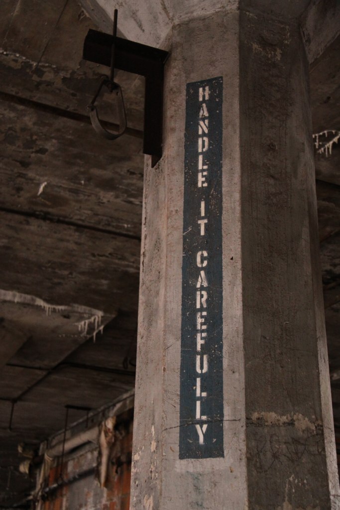 Weathered concrete pillar in a derelict industrial building stenciled vertically with "HANDLE IT CAREFULLY