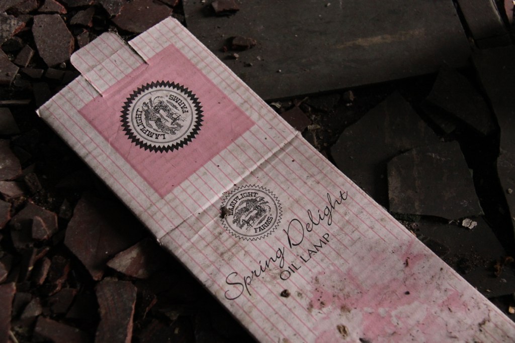 Discarded pink Lamplight Farms "Spring Delight Oil Lamp" box lying on dark rubble and debris.