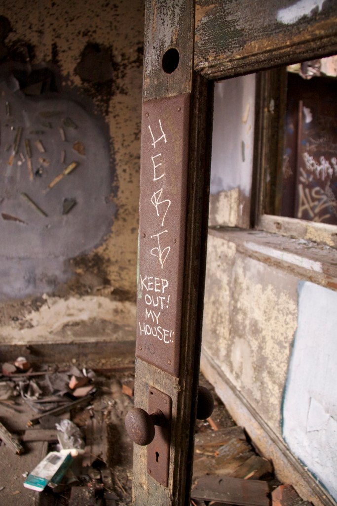 Weathered door in a decaying room, graffiti reading "HERB" and "KEEP OUT! MY HOUSE!" scrawled on its frame.