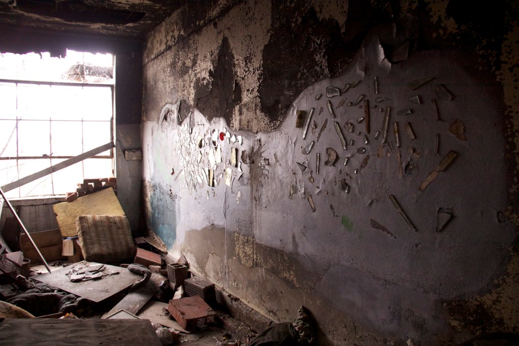 Debris and glass fragments embedded in the decaying wall of a fire-damaged, abandoned room with a broken window.