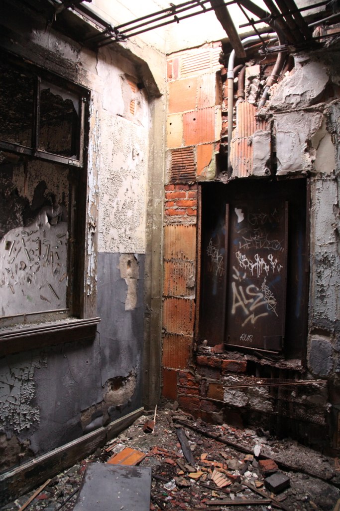 Decaying interior room with peeling walls, exposed brick, rubble-strewn floor, and a graffiti-tagged door reading "ALS.