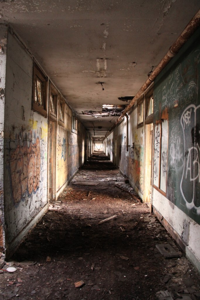 Long derelict hallway with graffiti-covered walls, boarded windows, and a debris-strewn floor receding into darkness.