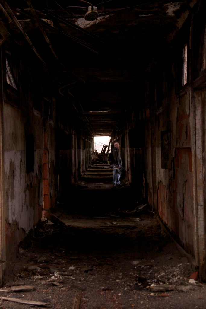 A lone figure stands in the distant light of a long, dark, decaying corridor strewn with debris.