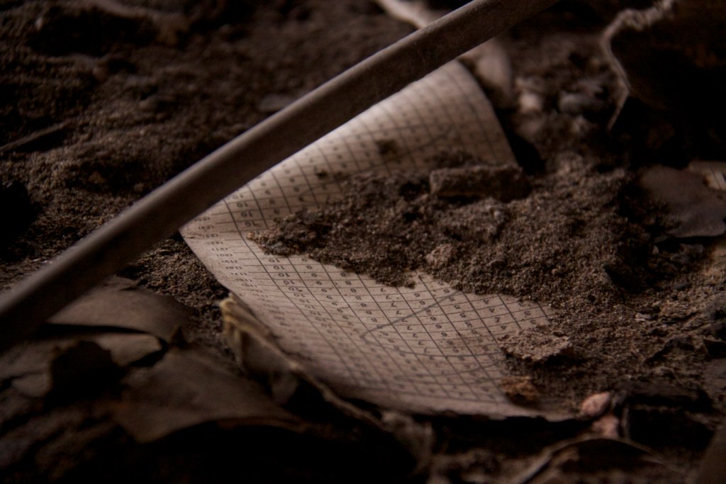Dusty numbered grid schedule ledger page half-buried in dirt and debris beneath a metal rod.