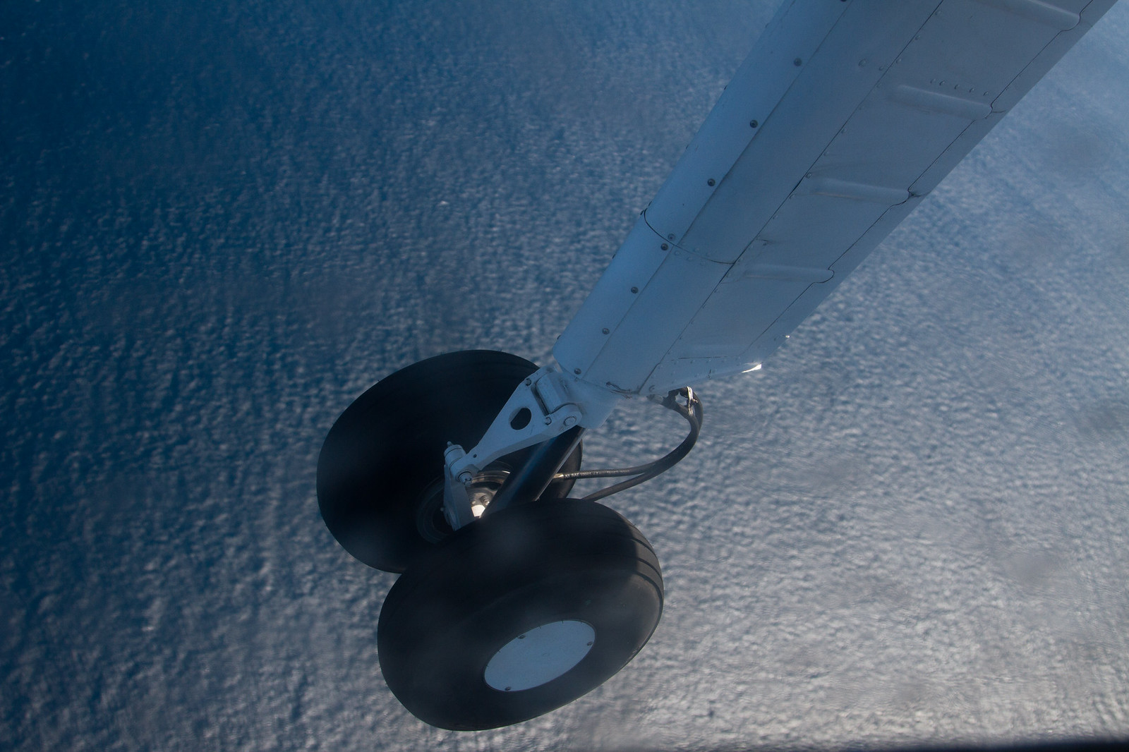 Close-up of a floatplane's landing wheel and white wing strut with the deep blue ocean far below.