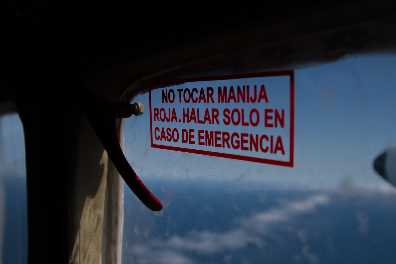 Small plane window with a red emergency-handle sign reading "NO TOCAR MANIJA ROJA," clouds below.