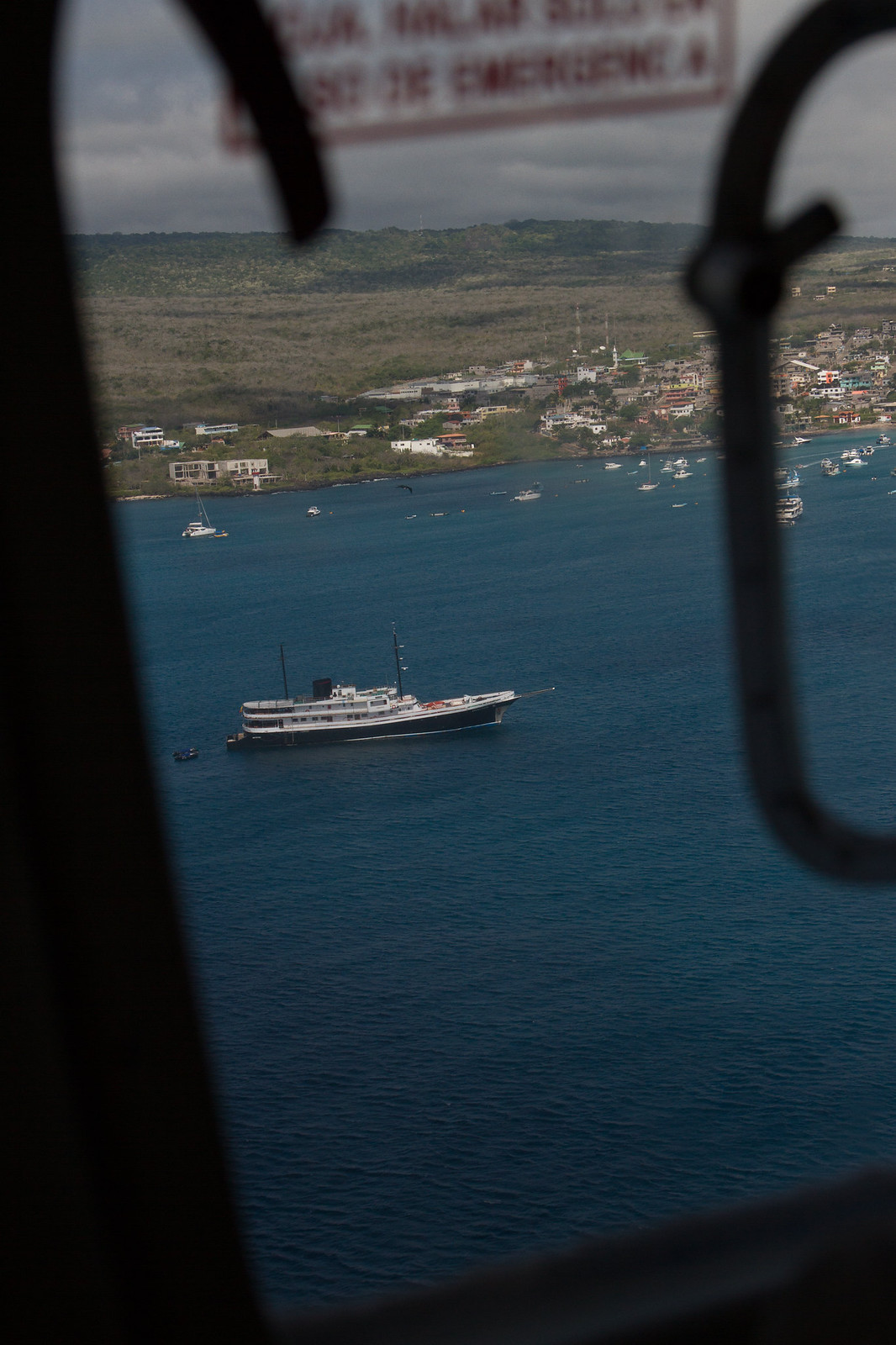 Aerial view through an aircraft window of boats anchored in a bay off a coastal Galápagos town.