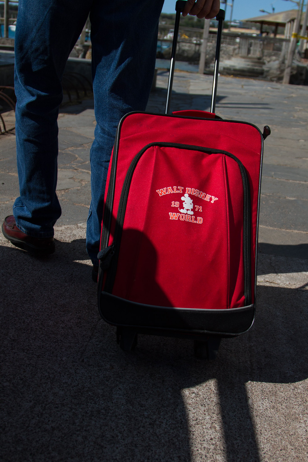 Person in jeans wheels a red "Walt Disney World 1971" rolling suitcase along a sunlit dock.