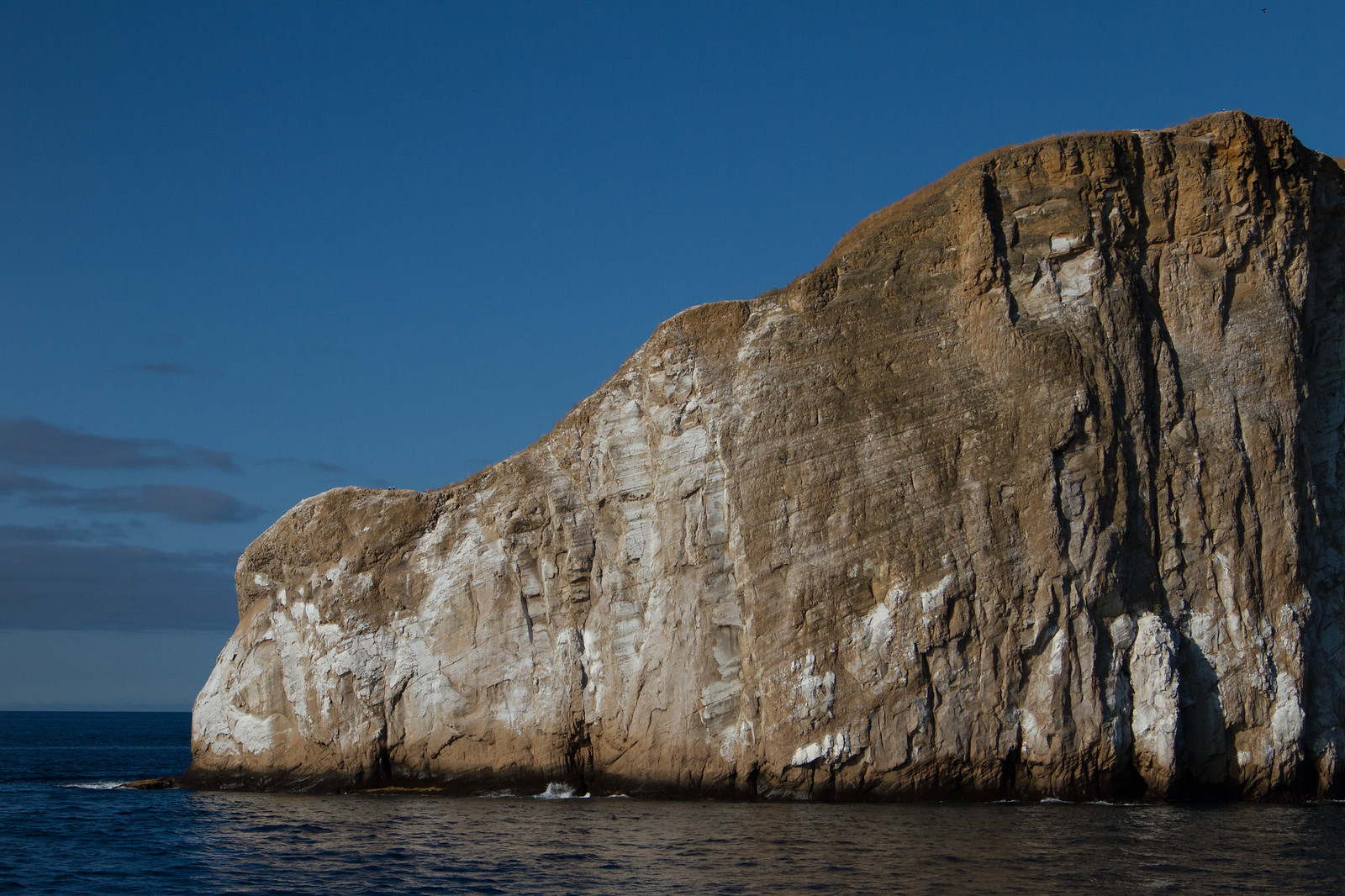 A rugged rocky cliff rising steeply from deep blue ocean under a clear evening sky.