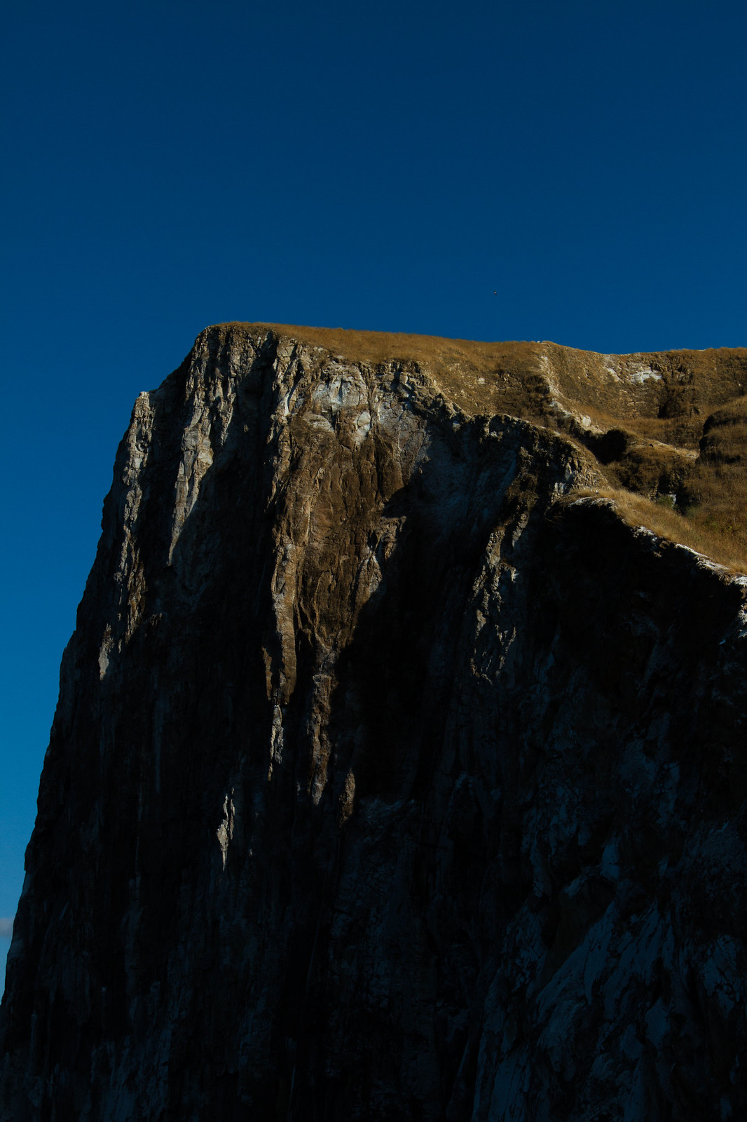 A rugged rocky cliff with a grassy top, lit by golden sunlight against a deep blue sky.
