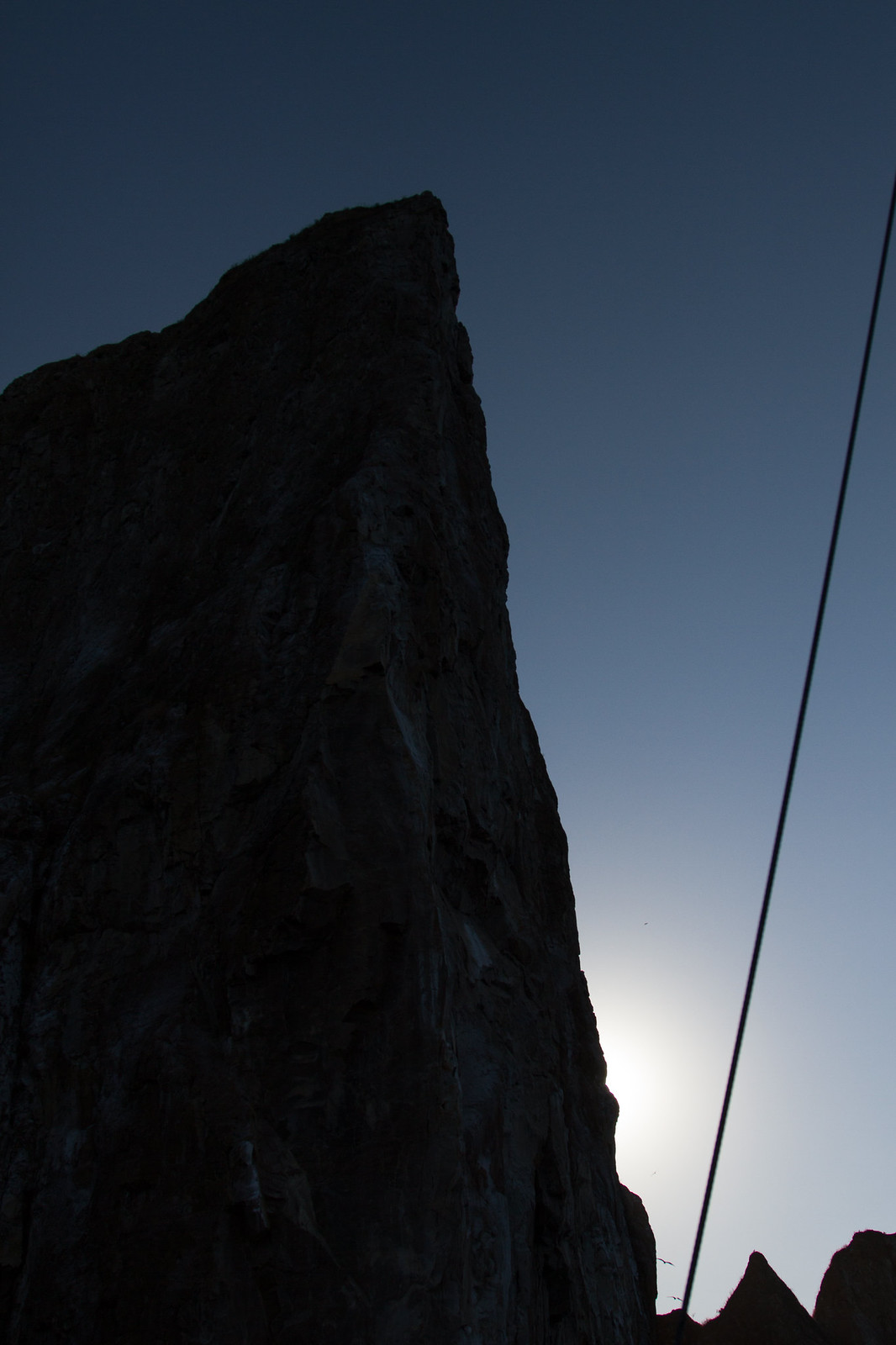 A towering rocky cliff silhouetted against a twilight sky with the sun glowing low behind it.