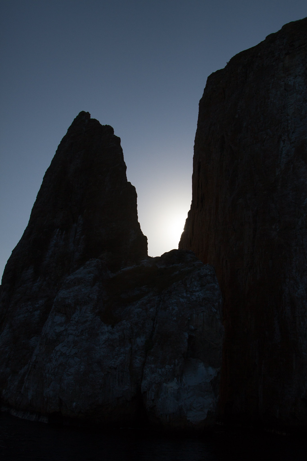 Silhouetted rock pinnacles of Kicker Rock at dusk with the setting sun glowing in the gap between them.