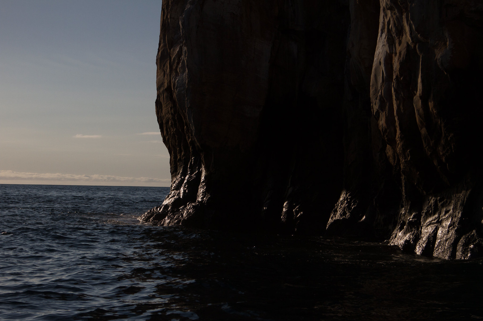 Steep dark sea cliff rising from calm ocean water beneath a pale sky at the horizon.