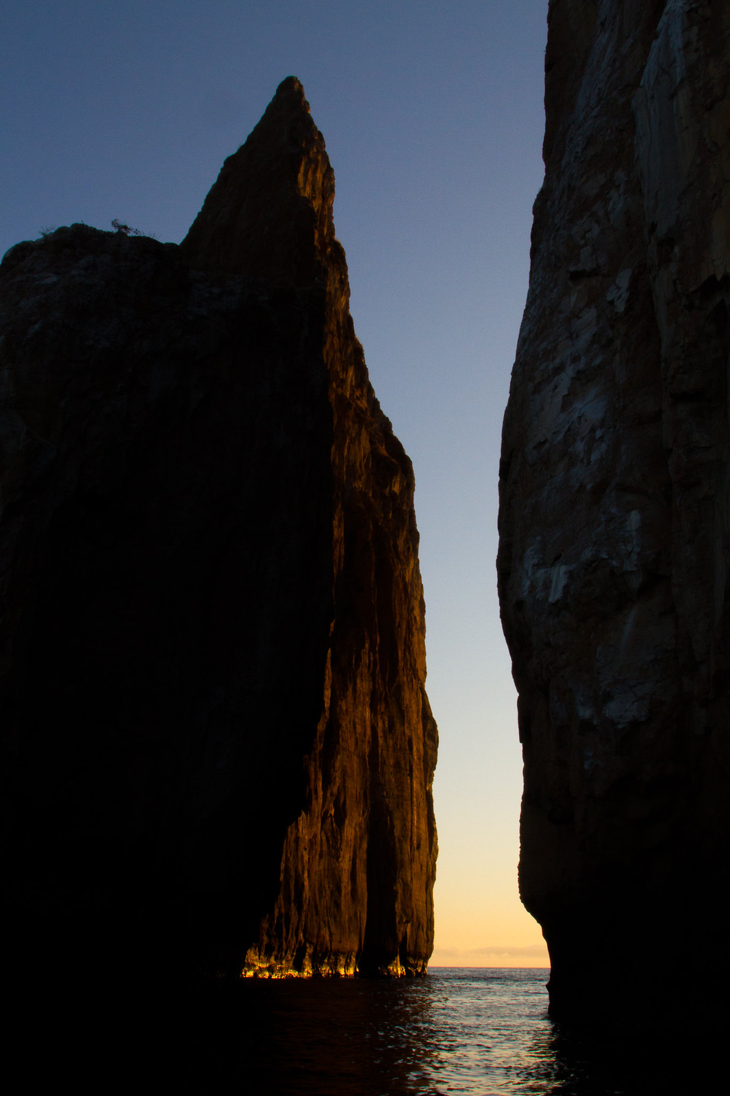 Sunlit rock pinnacle rising between dark sea cliffs at dusk over calm water, Galápagos.