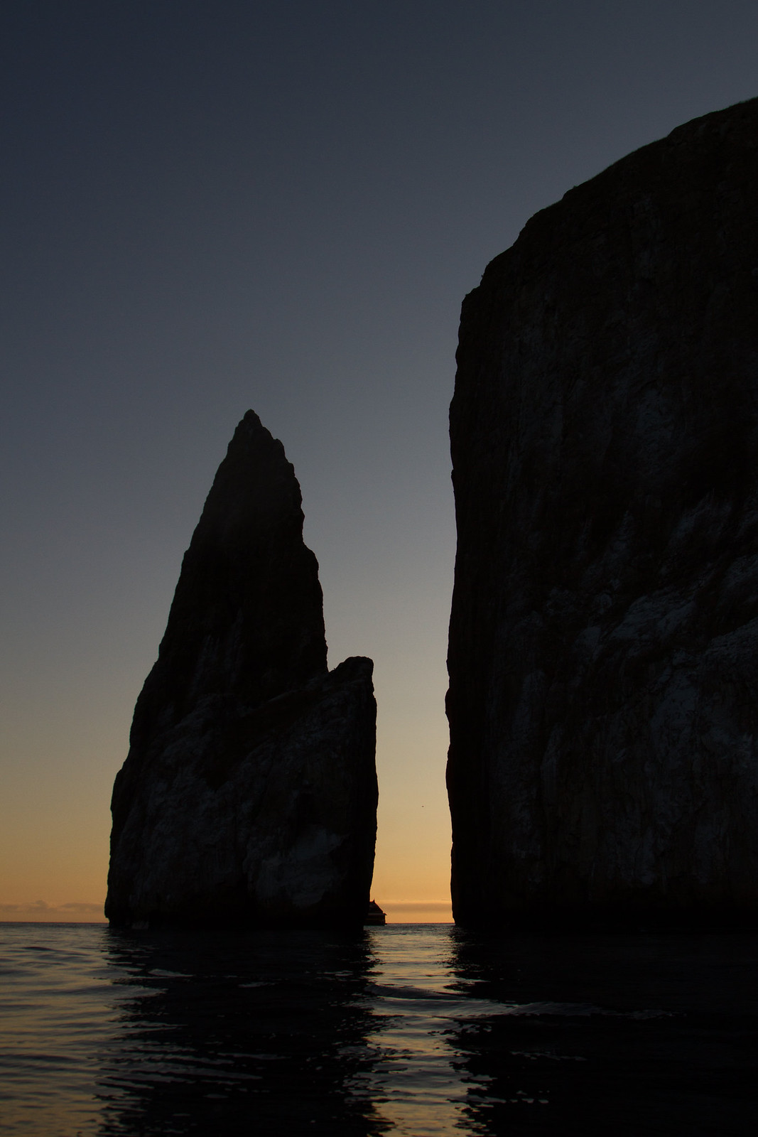 Kicker Rock's jagged sea cliffs silhouetted against a glowing orange sunset over calm ocean water.