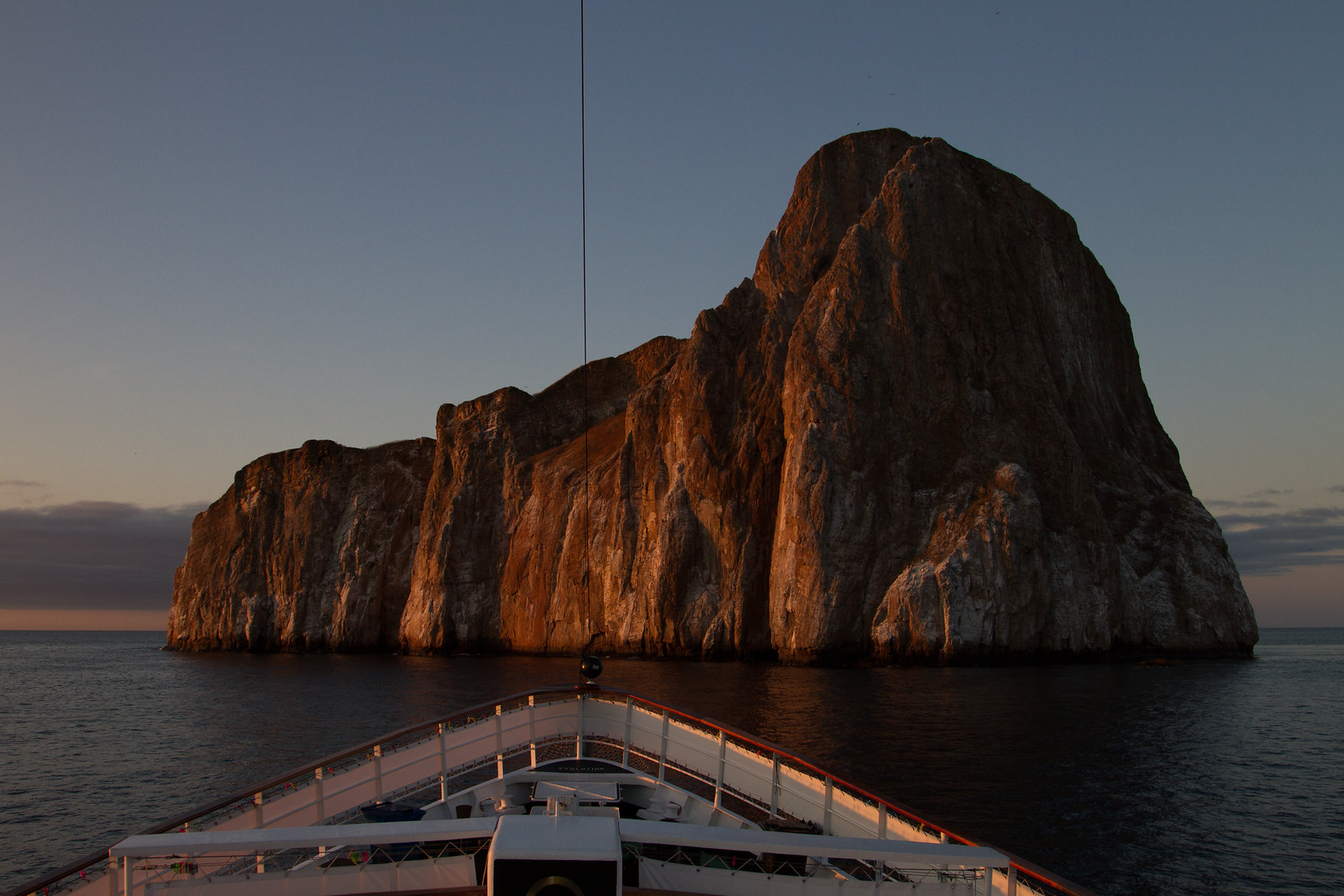A ship's bow faces the towering Kicker Rock cliffs glowing in Galápagos sunset light over calm sea.