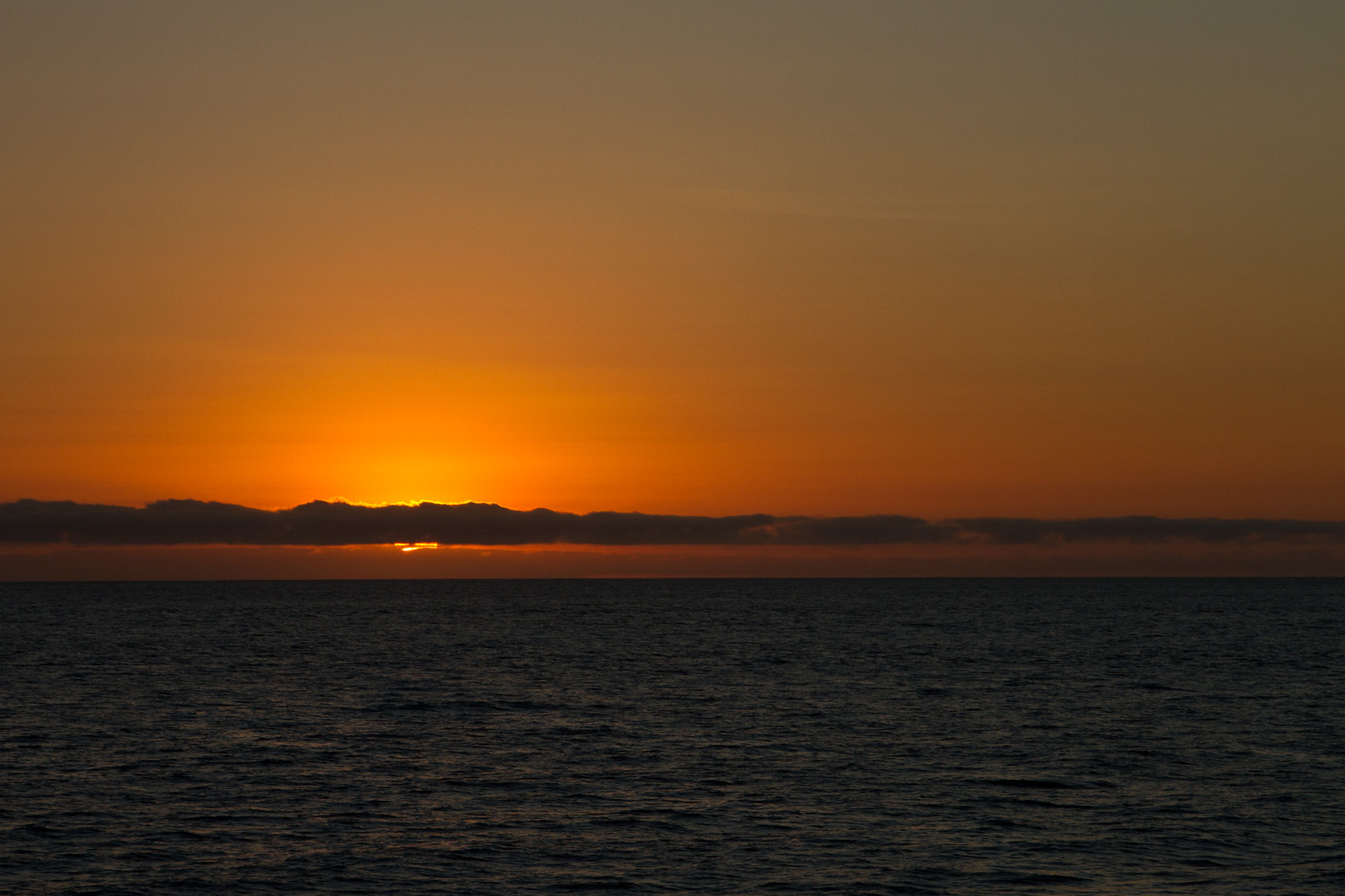 Fiery orange sun setting behind a low band of clouds over a calm, dark ocean horizon.