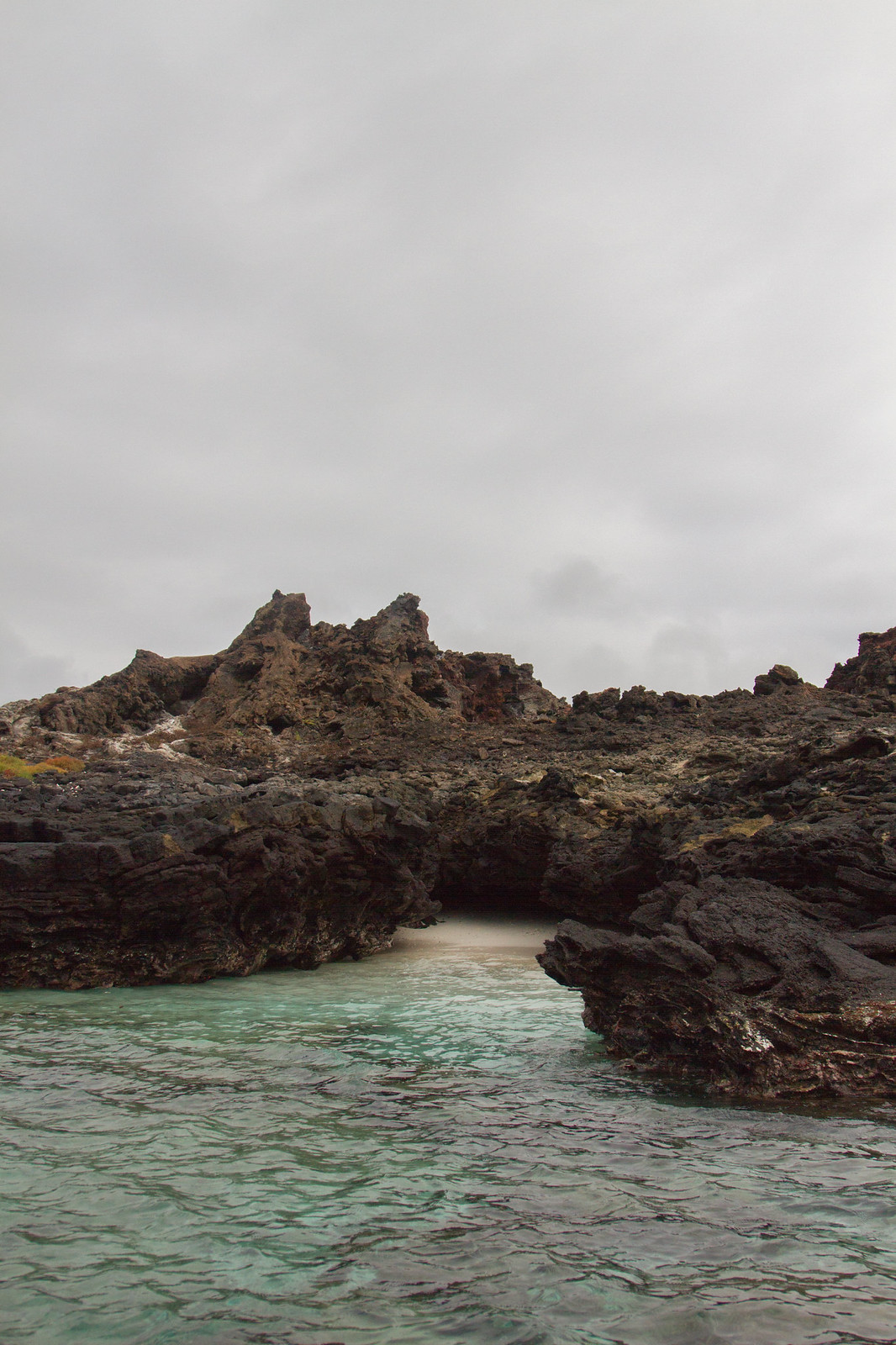 Rugged dark volcanic rock formations frame a hidden sandy cove of turquoise water under an overcast sky.